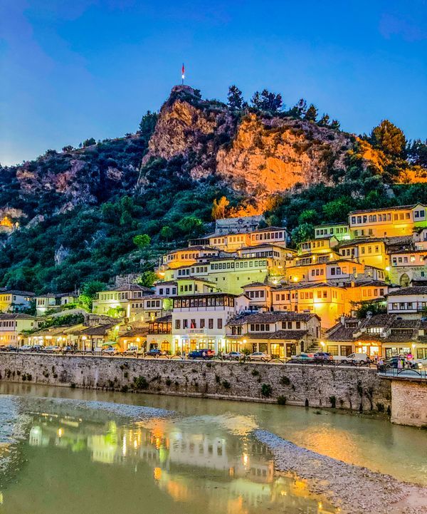 The 1000 windows of iconic Berat Albania. Evening lights from the city reflect on the river and the houses are painted green, yellow and orange. The blue hour highlights Berat Castle on the mountain.
