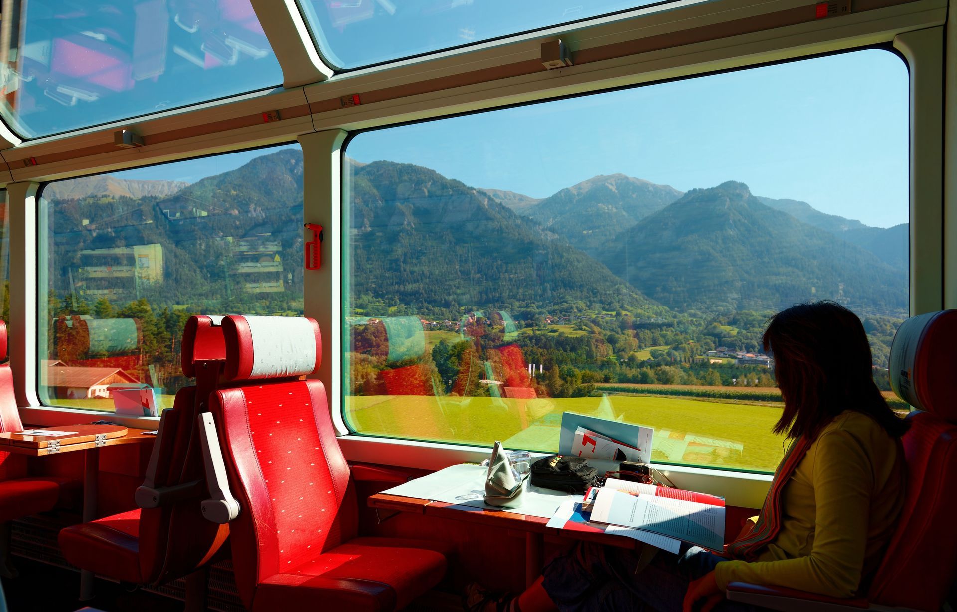 Tourists riding on Glacier Express, enjoying the idyllic scenery of Swiss countryside thru the wide panoramic windows, with sunlight cast thru the glass skylights on a sunny summer day in Switzerland